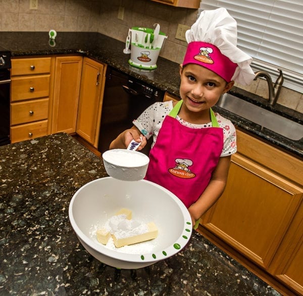 A young girl holding a measuring cup of flour over a bowl of flour and butter while smiling.